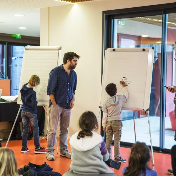 Daux enfants dessinent sur deux tableaux blancs accompanés par un dessinateur de BD